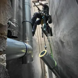 Pracownik na linach montuje rury wentylacyjne w wąskim szybie, widoczne elementy instalacji i fragment okna.