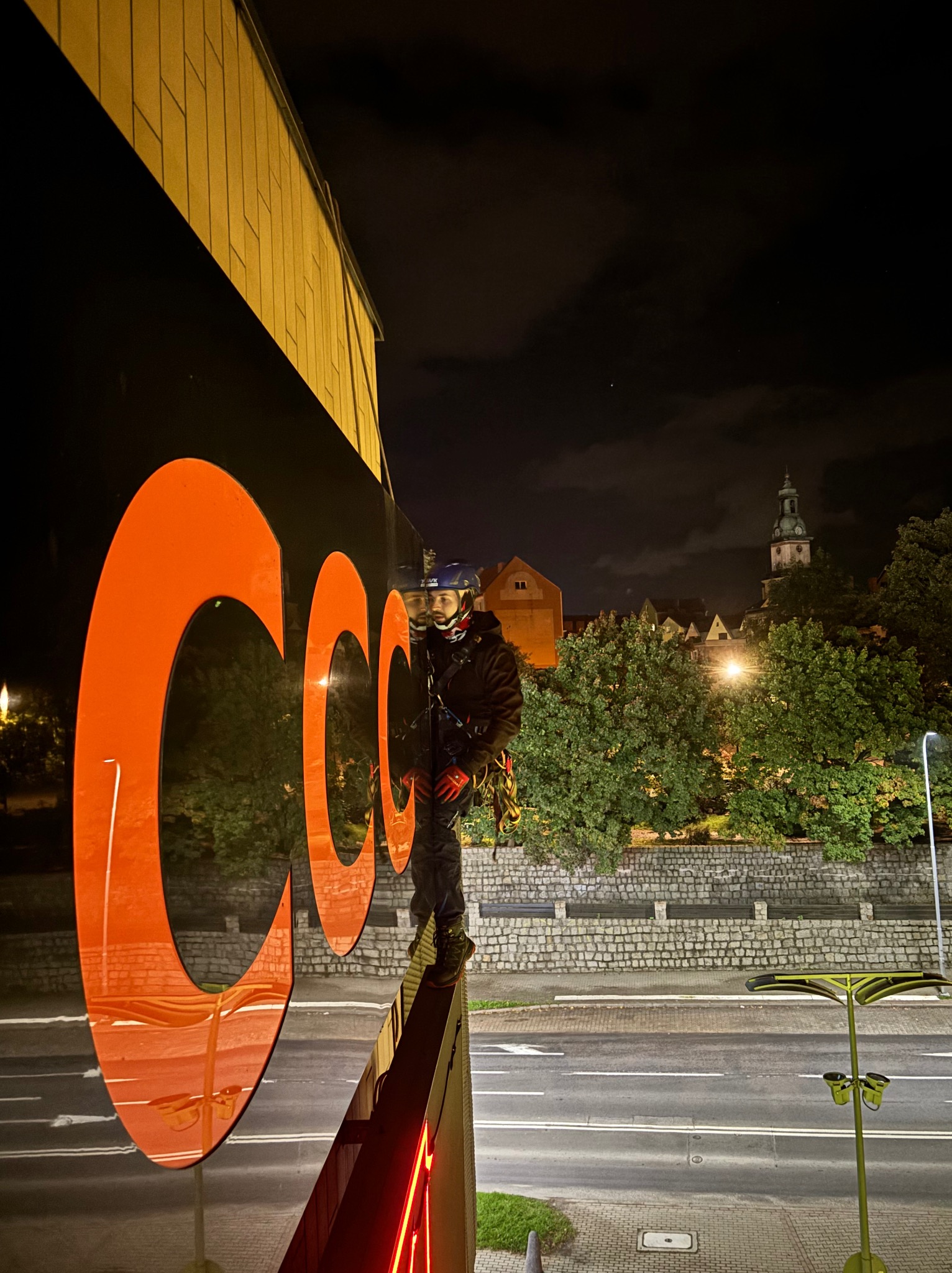 Pracownik w uprzęży, stojący na krawędzi budynku podczas nocnych prac na wysokości, z widocznym dużym, pomarańczowym logo na elewacji, w tle panorama miasta z wieżą kościelną.