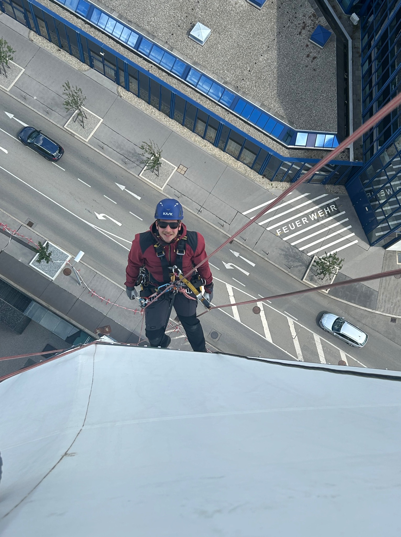 Pracownik w uprzęży linowej, w kasku i okularach przeciwsłonecznych, zwisa z białej elewacji budynku, widok z góry na ulicę z samochodami i napisem 'FEUERWEHR' na przejściu dla pieszych.