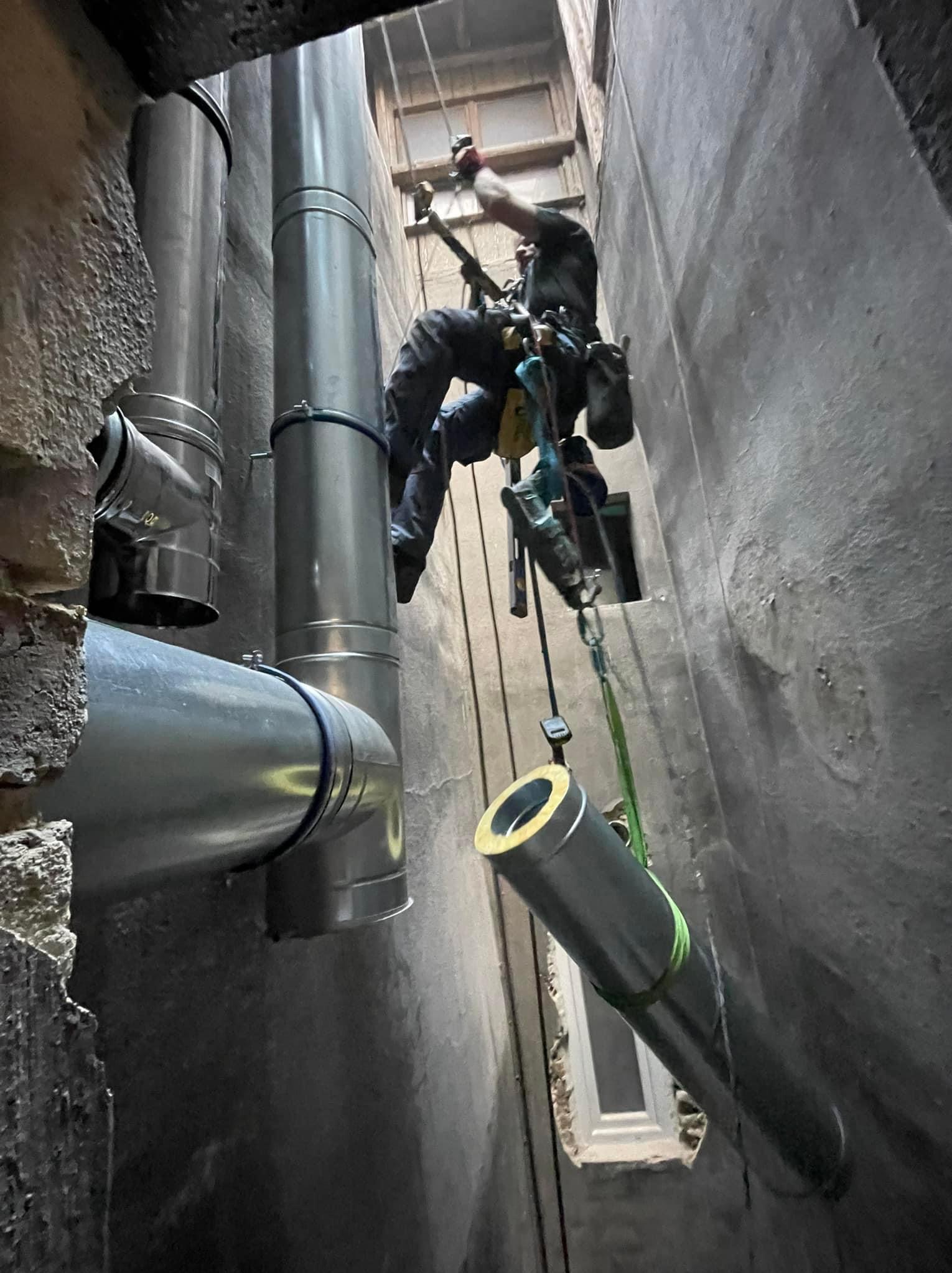 Pracownik na linach montuje rury wentylacyjne w wąskim szybie, widoczne elementy instalacji i fragment okna.