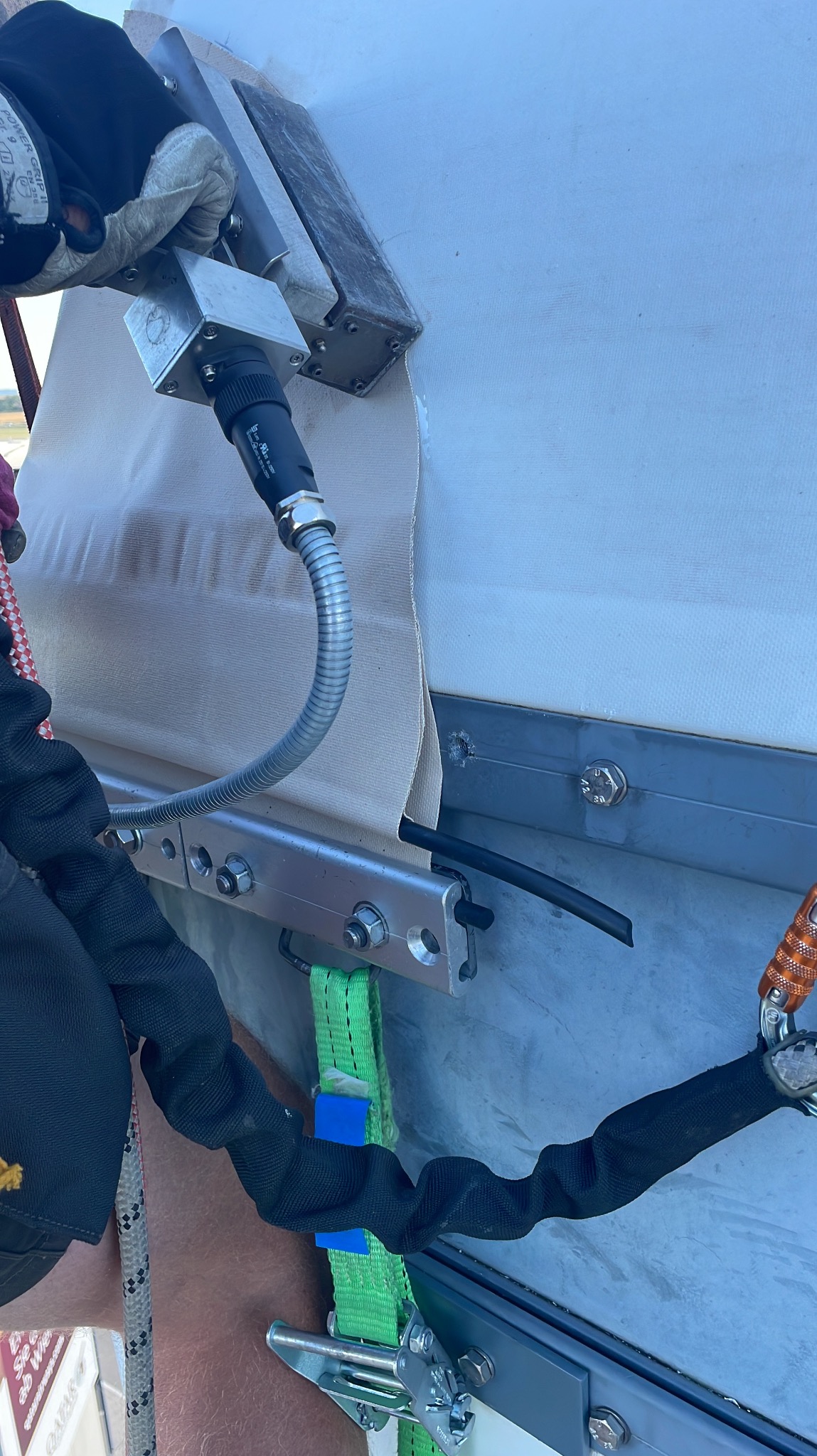 Praca na wysokości: Zgrzewanie plandeki na konstrukcji metalowej, widoczne zabezpieczenia linowe i narzędzie z kablem.