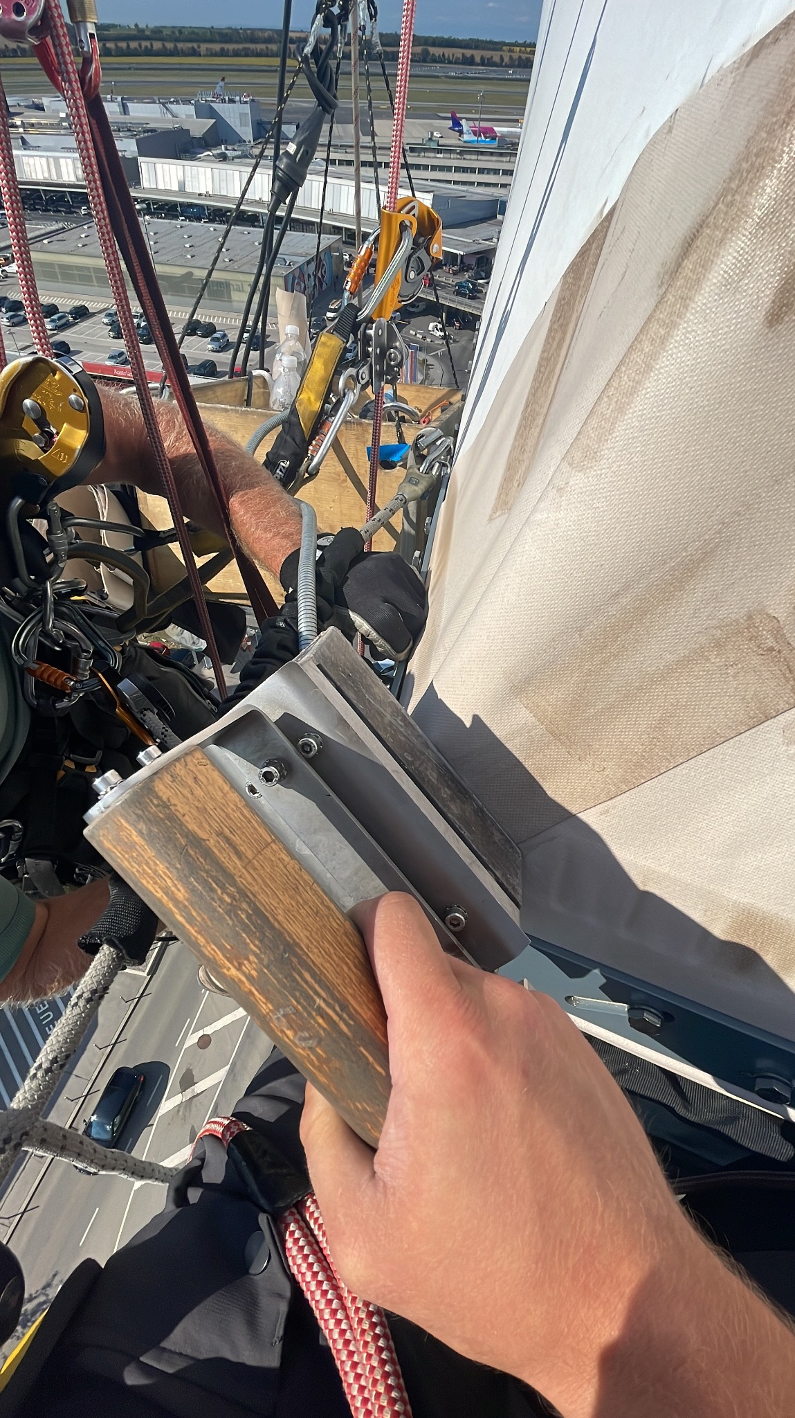 Pracownik w uprzęży, z bliska trzymający narzędzie do czyszczenia pionowej powierzchni, prawdopodobnie elewacji budynku. W tle widoczny port lotniczy z samolotami.