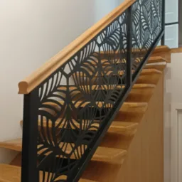 Drewniane schody z czarną, ażurową barierką o motywie liści, wykończoną drewnianą poręczą. Nowoczesne oświetlenie i dekoracje ścienne w tle.