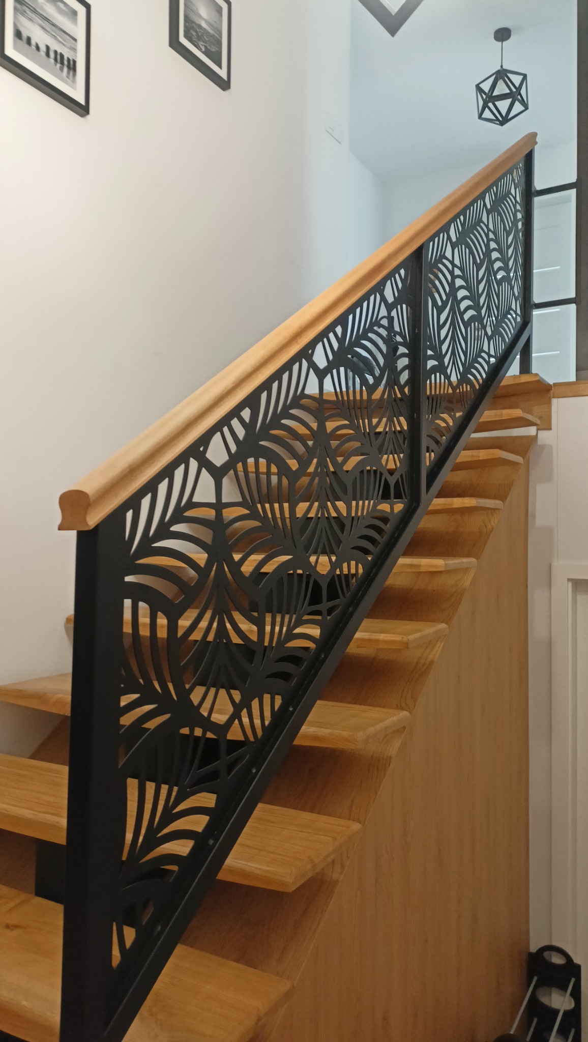 Drewniane schody z czarną, ażurową barierką o motywie liści, wykończoną drewnianą poręczą. Nowoczesne oświetlenie i dekoracje ścienne w tle.