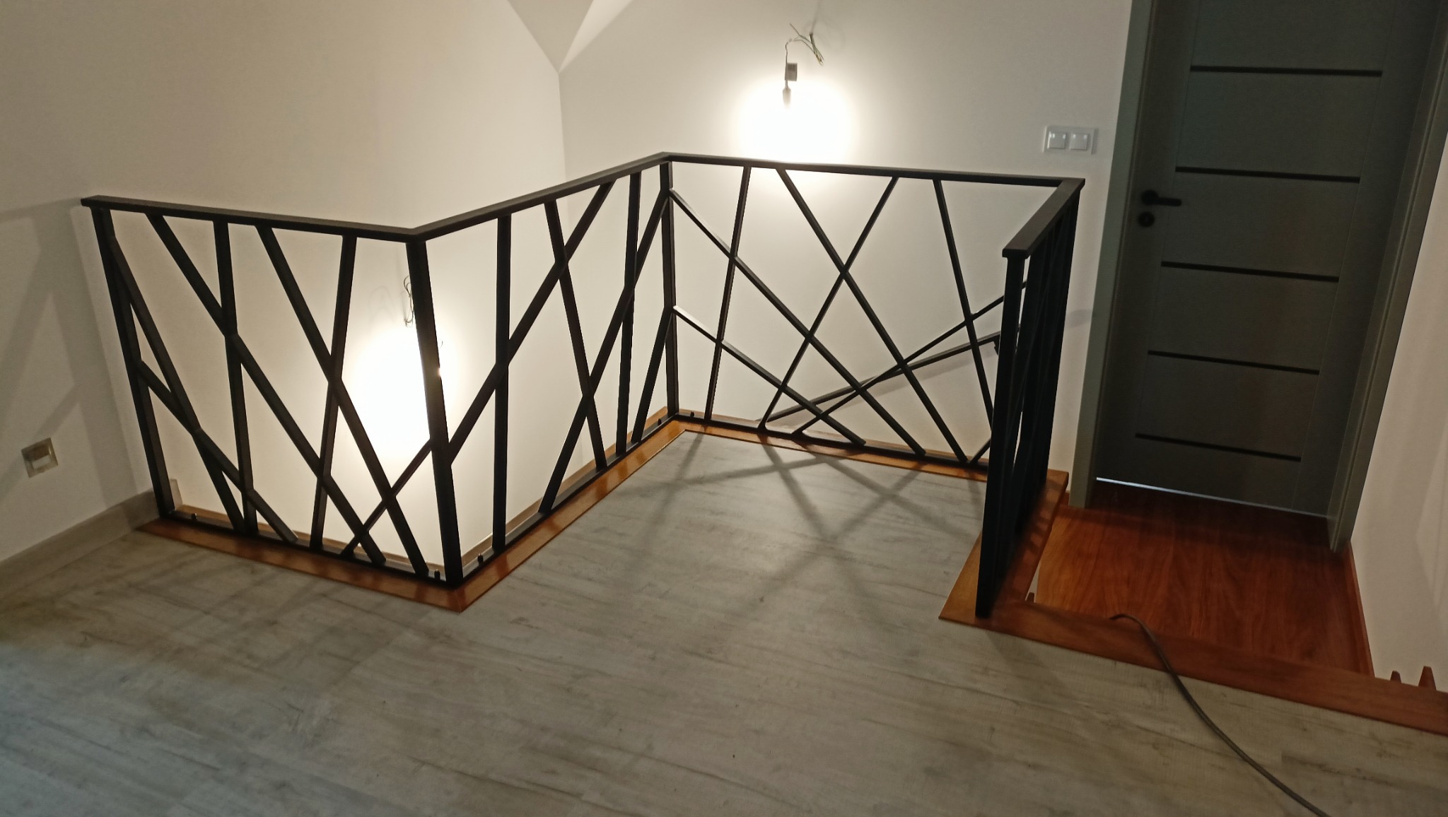 Nowoczesna, czarna balustrada z geometrycznym wzorem na jasnym tle ściany i podłogi, widoczna z góry, z fragmentem drzwi w tle.