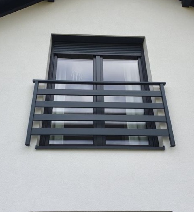Nowoczesna, szara balustrada okienna z poziomych profili, zamontowana na białej elewacji budynku. Okno z roletą zewnętrzną, ujęcie od dołu.