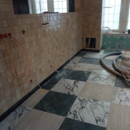 Adbud - Ściany wyłożone drobną, beżową mozaiką z widocznymi otworami na instalacje, podłoga w czarno-białą szachownicę z marmurowych płyt, częściowo zakryta wężem odkurzacza i wiadrem z farbą, okno w tle.