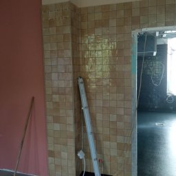 Adbud - Fragment wnętrza w trakcie prac wykończeniowych: ściana z brązowych płytek, widoczne listwy dystansowe, obok ściana pomalowana na różowo, na podłodze sprzęt i oświetlenie robocze, w tle widoczna...