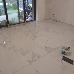 Adbud - Jasne, marmurowe płytki podłogowe w trakcie układania, widoczne narzędzia glazurnicze: paca i pojemnik z klejem. W tle otwarte drzwi balkonowe i fragment ogrodu.
