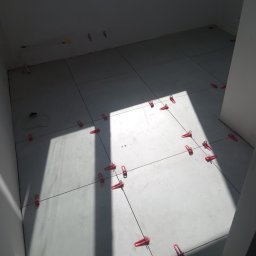 Adbud - Świeżo ułożone, jasnoszare płytki podłogowe w trakcie poziomowania za pomocą czerwonych klipsów i krzyżyków dystansowych, widoczne kable na ścianie, silne światło dzienne padające z góry.