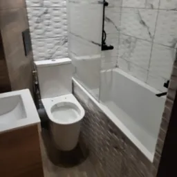 Nowoczesna łazienka po renowacji: biała armatura, wanna z czarną armaturą i szklanym parawanem, umywalka z drewnianą szafką, płytki imitujące drewno na podłodze i strukturalne białe płytki...