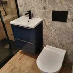 Nowoczesna łazienka z wiszącą toaletą Roca, szafką umywalkową w kolorze granatowym z czarną baterią, szklaną kabiną prysznicową oraz płytkami imitującymi lastryko na ścianach.