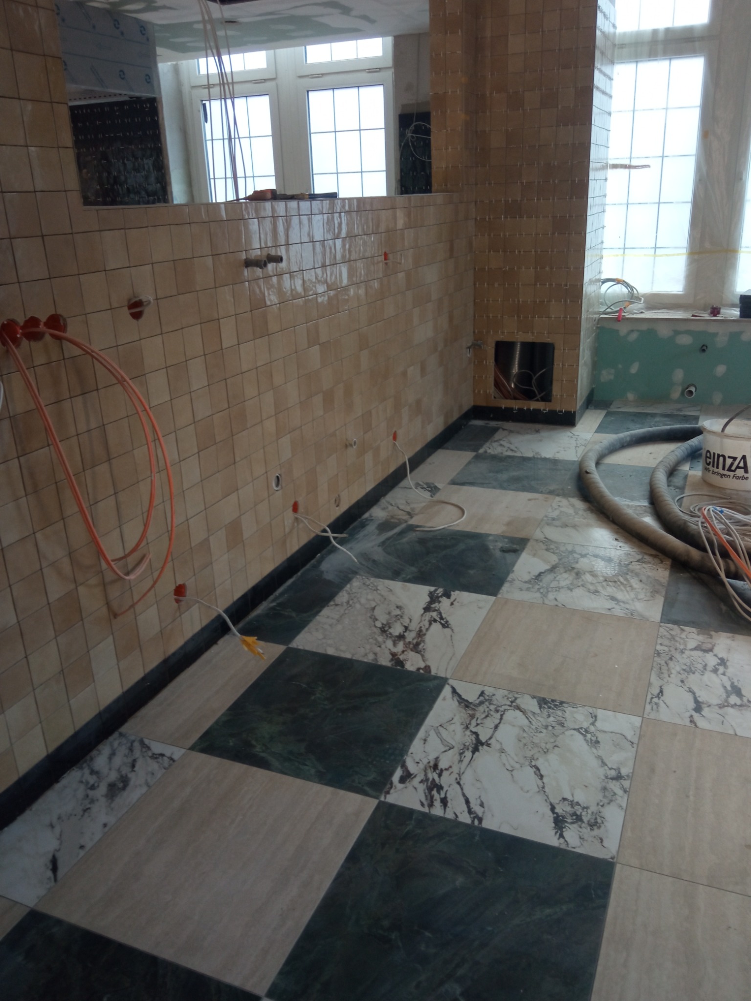 Ściany wyłożone drobną, beżową mozaiką z widocznymi otworami na instalacje, podłoga w czarno-białą szachownicę z marmurowych płyt, częściowo zakryta wężem odkurzacza i wiadrem z farbą, okno w tle.