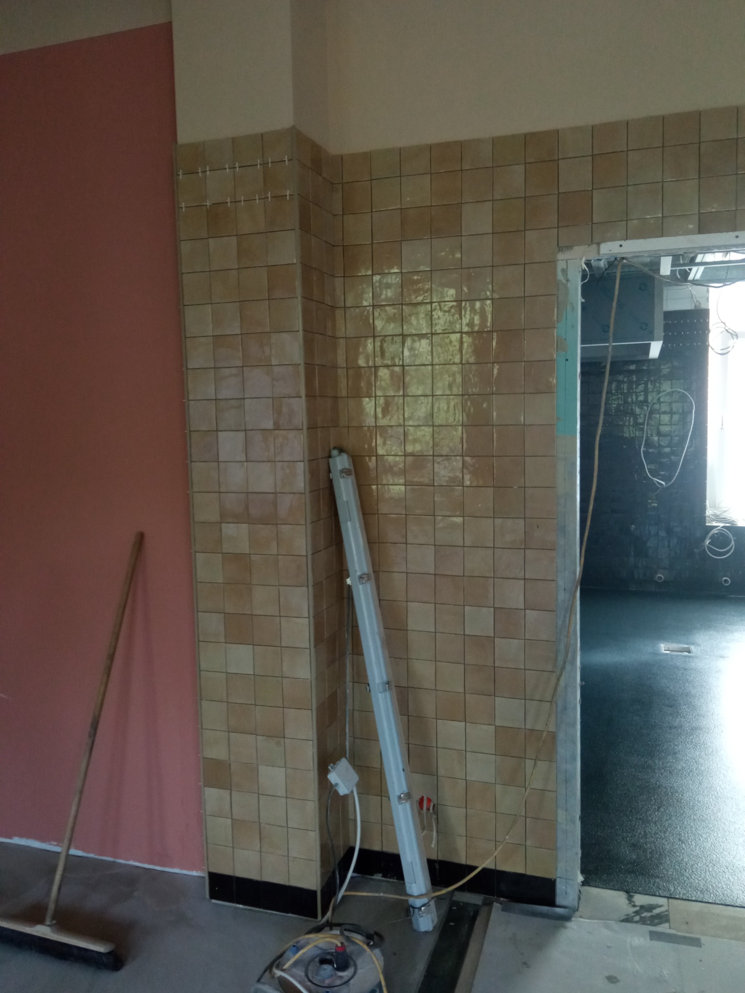 Fragment wnętrza w trakcie prac wykończeniowych: ściana z brązowych płytek, widoczne listwy dystansowe, obok ściana pomalowana na różowo, na podłodze sprzęt i oświetlenie robocze, w tle widoczna...