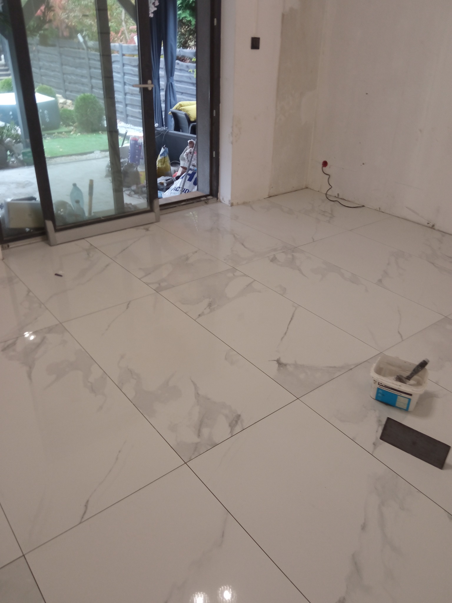 Jasne, marmurowe płytki podłogowe w trakcie układania, widoczne narzędzia glazurnicze: paca i pojemnik z klejem. W tle otwarte drzwi balkonowe i fragment ogrodu.