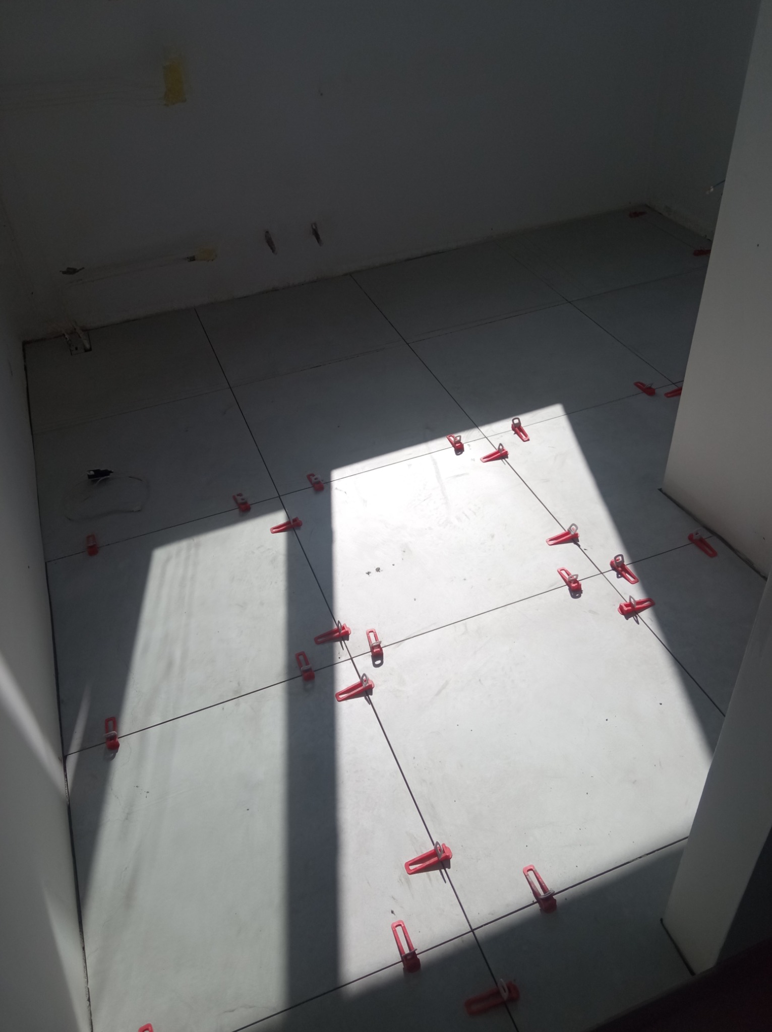 Świeżo ułożone, jasnoszare płytki podłogowe w trakcie poziomowania za pomocą czerwonych klipsów i krzyżyków dystansowych, widoczne kable na ścianie, silne światło dzienne padające z góry.