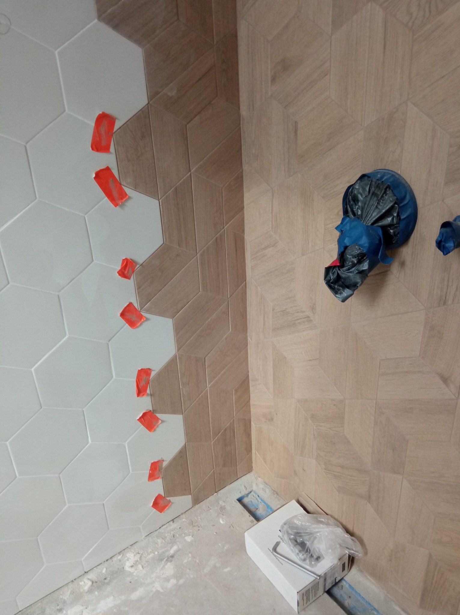 Ściana w trakcie układania płytek: heksagonalne białe płytki sąsiadują z drewnopodobnymi płytkami o wzorze 3D, zabezpieczone pomarańczową taśmą. Na podłodze widoczny biały karton i zabezpieczone rury.