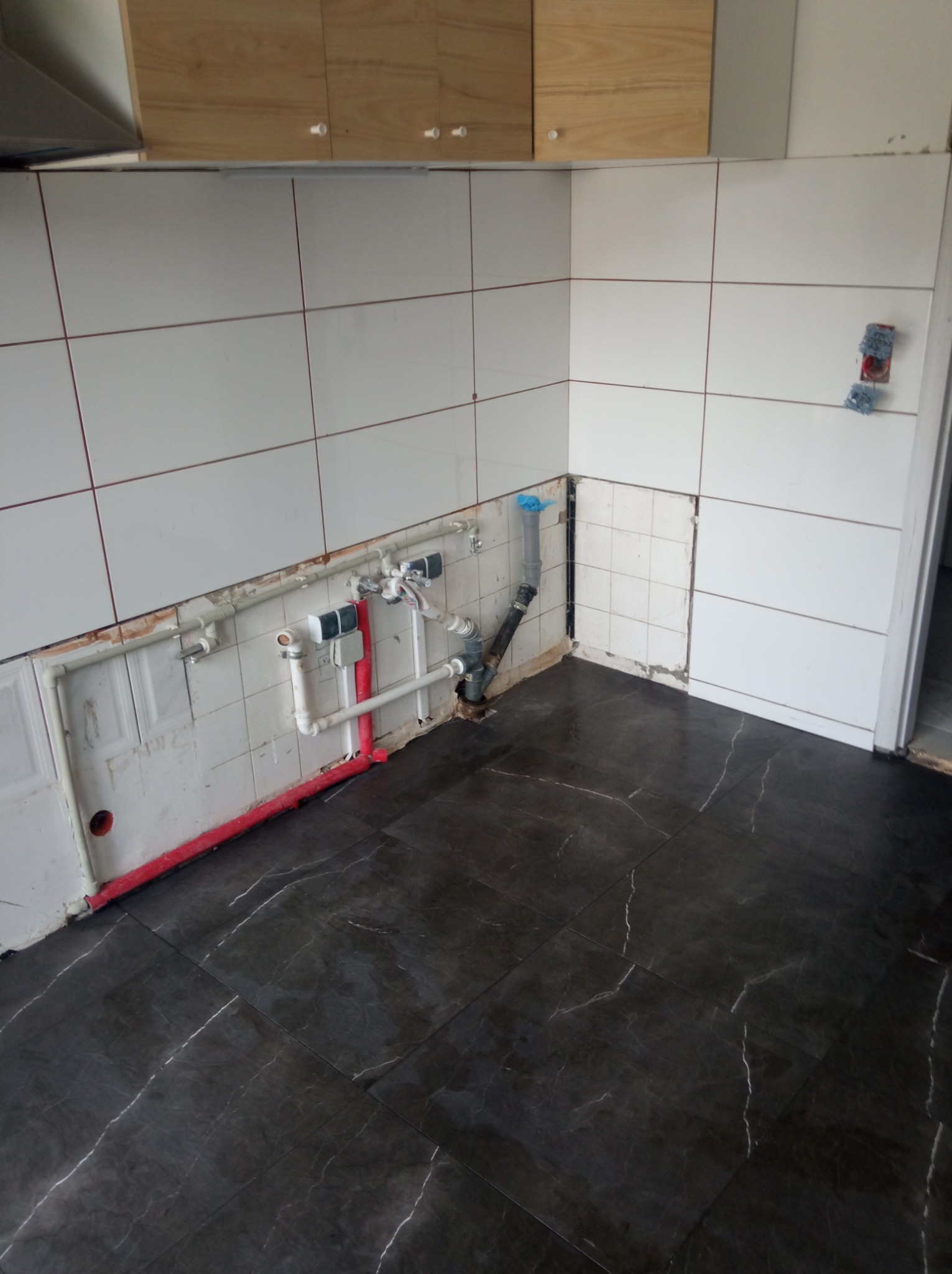 Częściowo wykończona kuchnia z ciemnymi płytkami podłogowymi imitującymi marmur i białymi płytkami ściennymi z bordową fugą, widoczne rury instalacji wodnej i fragmenty niezabezpieczonych ścian.