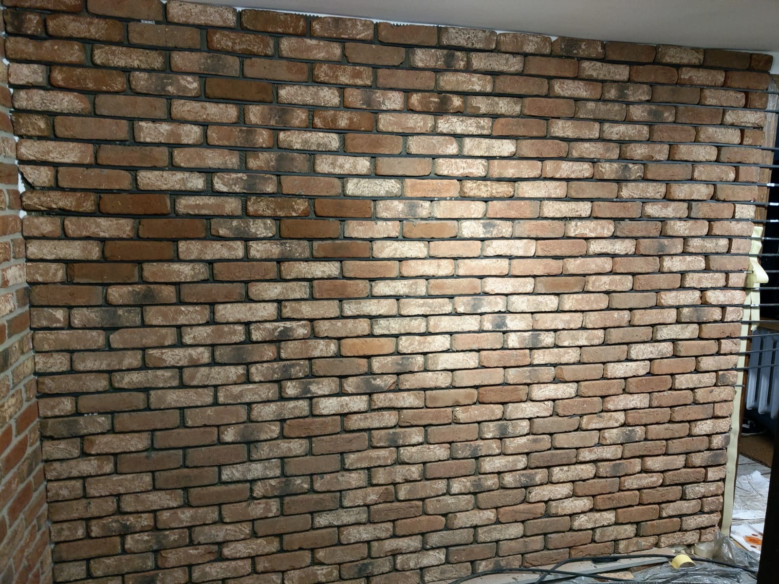 Ściana wyłożona dekoracyjną cegłą w odcieniach brązu i beżu, z ciemną fugą, widoczne niedoskonałości w ułożeniu i odcieniach cegieł, w tle fragment grzejnika i podłogi zabezpieczonej folią.