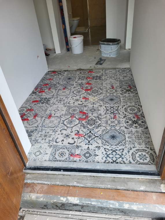 Układanie wzorzystych, szaro-białych płytek podłogowych w korytarzu z użyciem czerwonych krzyżyków dystansowych, widoczne wiaderka i narzędzia w tle.