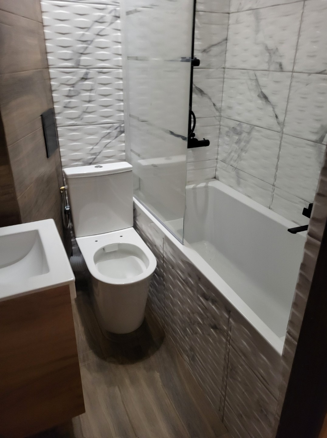 Nowoczesna łazienka po renowacji: biała armatura, wanna z czarną armaturą i szklanym parawanem, umywalka z drewnianą szafką, płytki imitujące drewno na podłodze i strukturalne białe płytki...