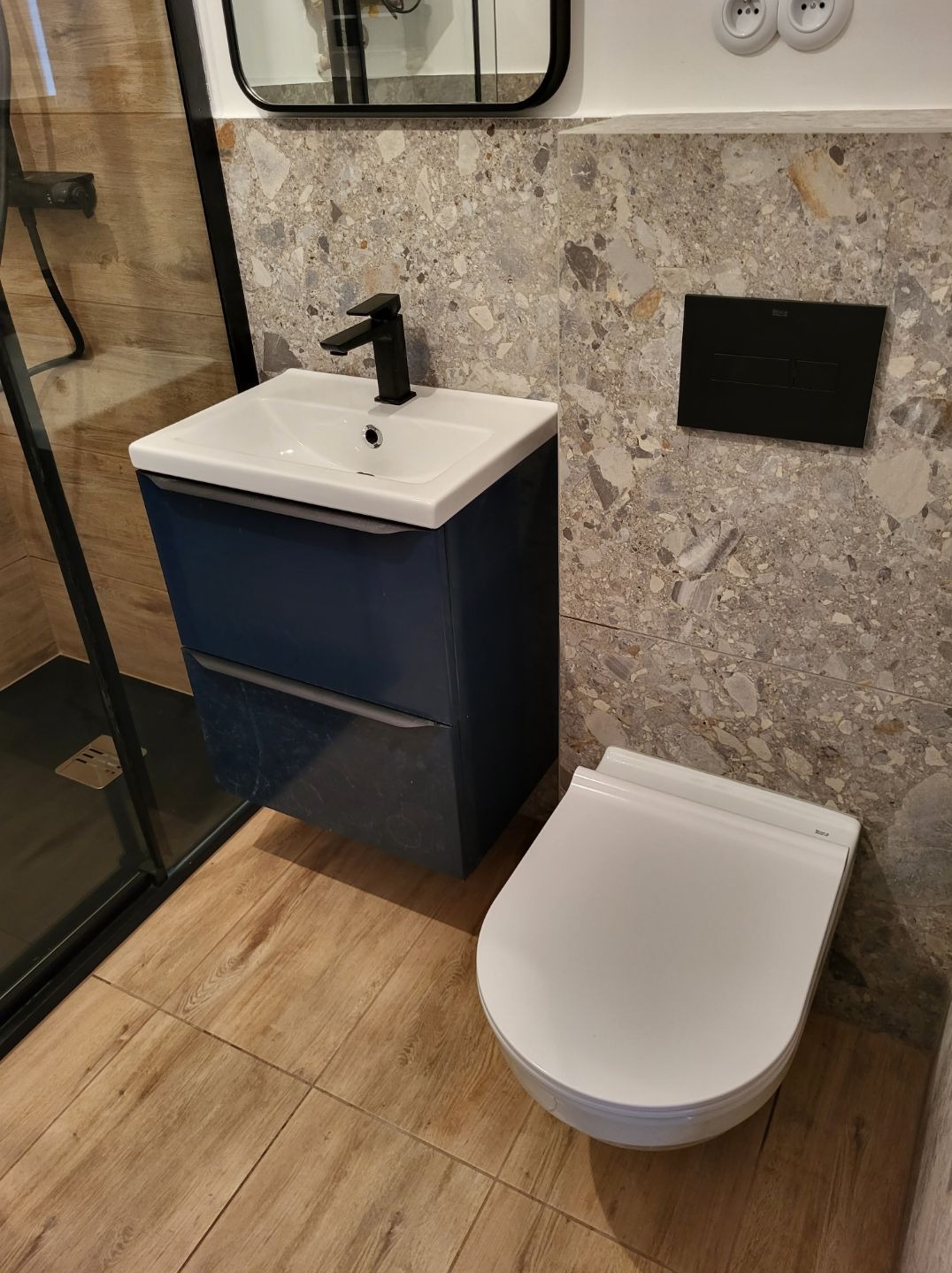 Nowoczesna łazienka z wiszącą toaletą Roca, szafką umywalkową w kolorze granatowym z czarną baterią, szklaną kabiną prysznicową oraz płytkami imitującymi lastryko na ścianach.