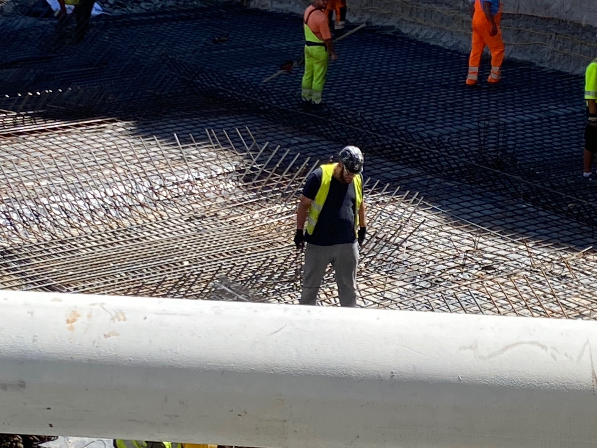 Pracownicy w kamizelkach układają siatkę zbrojeniową na budowie. Widoczne elementy konstrukcji żelbetowej i narzędzia. Praca zespołowa na placu budowy.