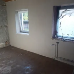 Pokój w trakcie prac wykończeniowych: ściany po szpachlowaniu, jedno okno z nową stolarką, drugie zakryte folią, widoczne rury instalacji grzewczej przy podłodze.