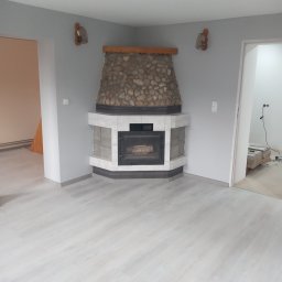 Bolesław - Wykończony kominek narożny z obudową z kamienia naturalnego i białych płytek w szarym pomieszczeniu z panelami podłogowymi, widoczne elementy dekoracyjne na ścianie oraz prace wykończeniowe...