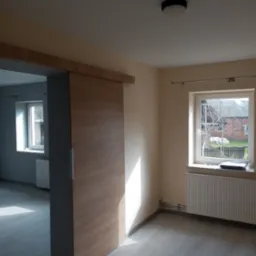 Wnętrze pokoju po remoncie z przesuwnymi drzwiami z okleiną drewnopodobną, grzejnikiem pod oknem i świeżo pomalowanymi ścianami w kolorze kremowym.