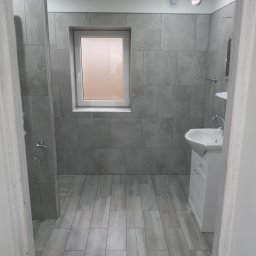 Bolesław - Wykończona łazienka w szarościach: płytki imitujące kamień na ścianach i podłodze, biała umywalka z szafką, okno z rozmytym widokiem, minimalistyczne oświetlenie sufitowe.