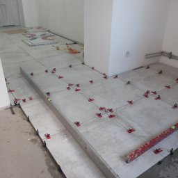 Bolesław - Układanie szarych płytek podłogowych z użyciem systemu poziomowania płytek, widoczna poziomica i puszki elektryczne w ścianie.