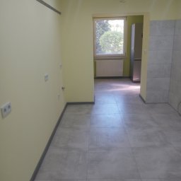 Bolesław - Wnętrze w trakcie remontu: żółte ściany z widocznymi gniazdkami elektrycznymi, szare płytki podłogowe, listwa montażowa pod sufitem i częściowo wykończona ściana z płytek w tle.