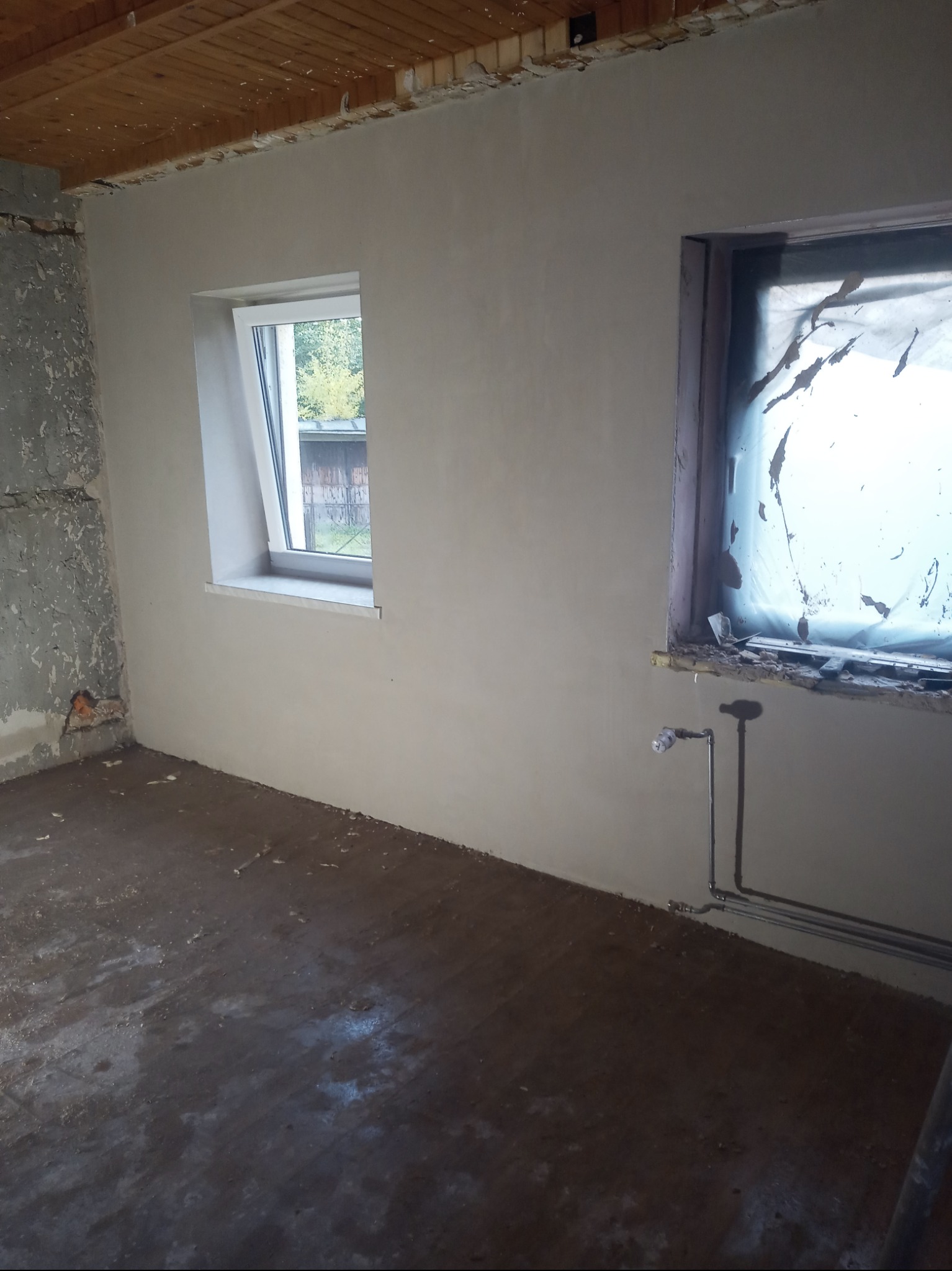 Pokój w trakcie prac wykończeniowych: ściany po szpachlowaniu, jedno okno z nową stolarką, drugie zakryte folią, widoczne rury instalacji grzewczej przy podłodze.