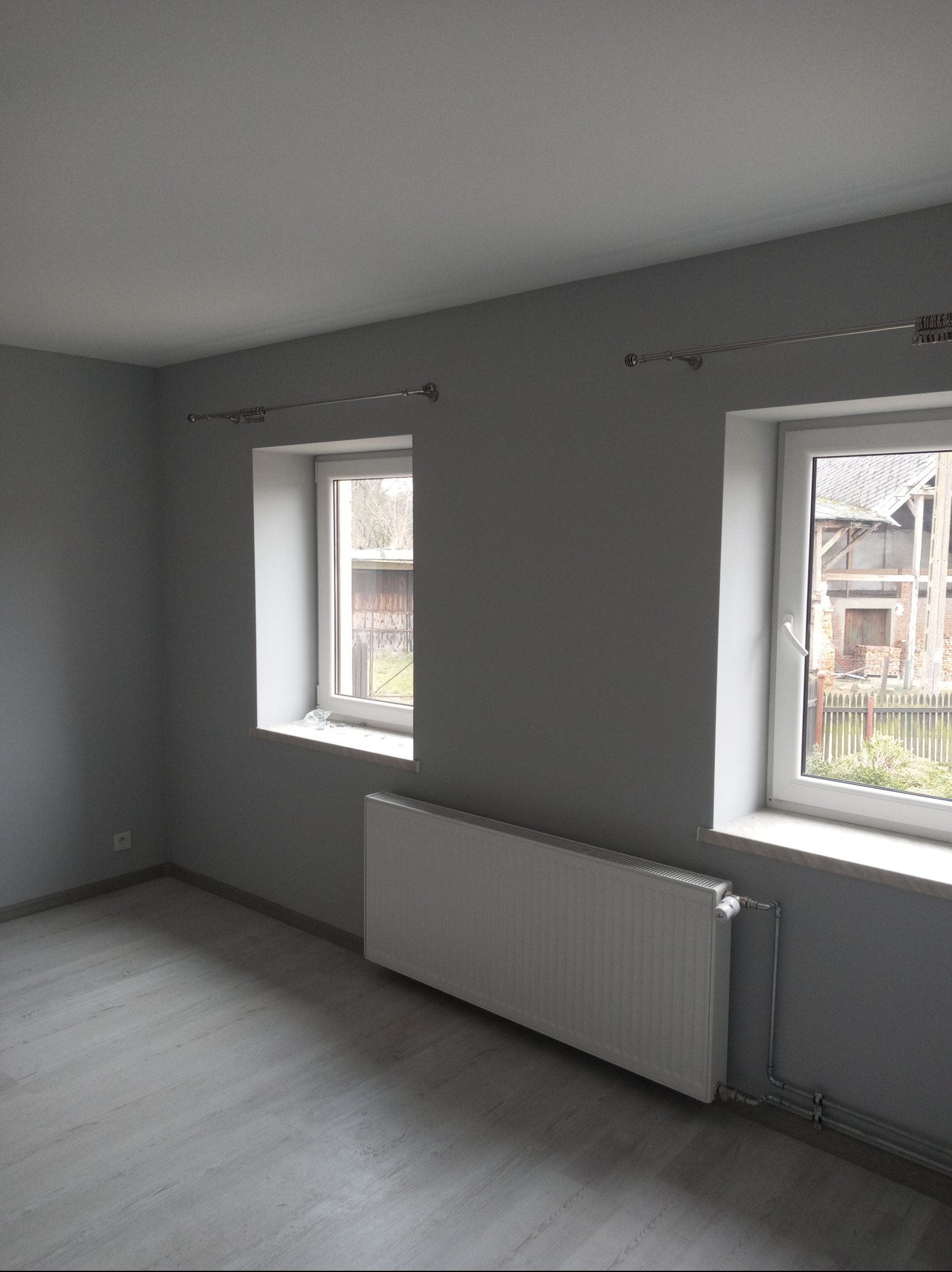 Pomalowane na szaro wnętrze pokoju z dwoma oknami, grzejnikiem i jasną podłogą z paneli, widok na zewnątrz z elementami wiejskiej zabudowy.