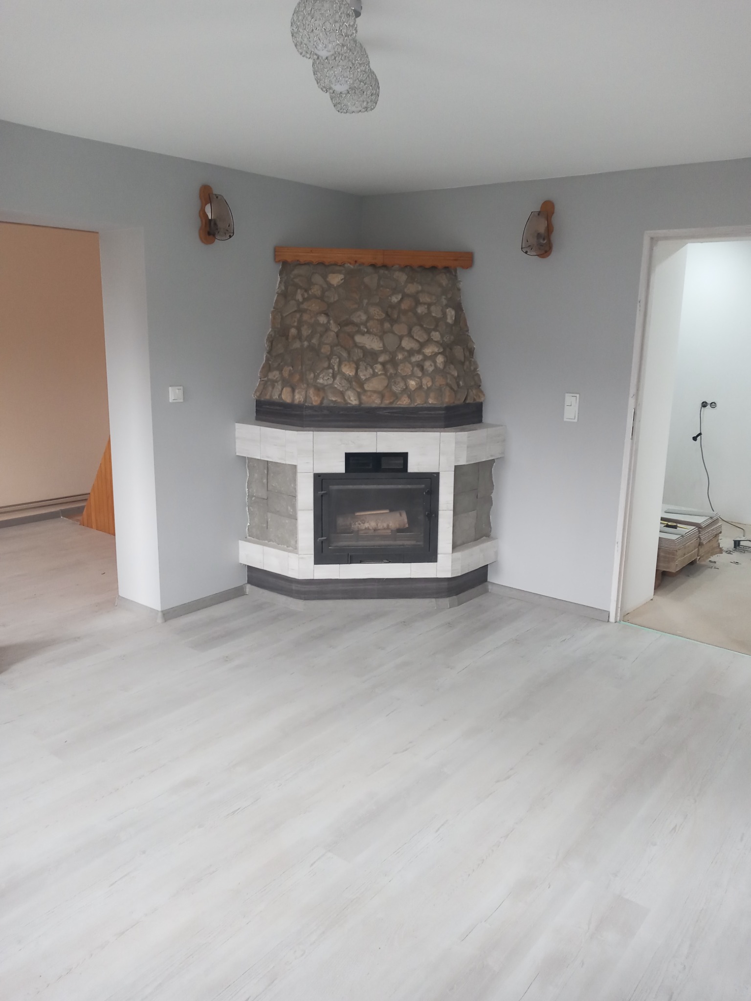 Wykończony kominek narożny z obudową z kamienia naturalnego i białych płytek w szarym pomieszczeniu z panelami podłogowymi, widoczne elementy dekoracyjne na ścianie oraz prace wykończeniowe...