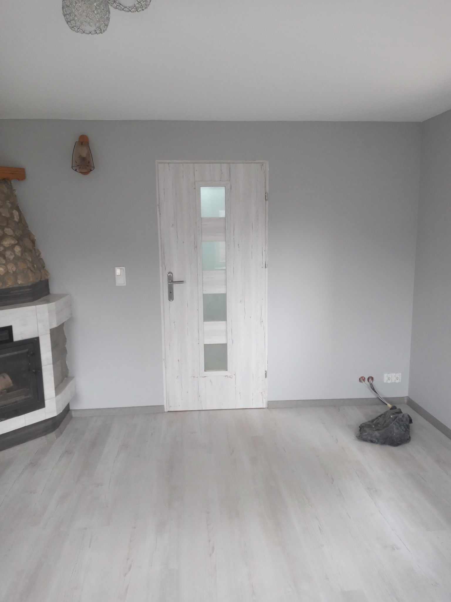 Pusty pokój z szarymi ścianami, kominkiem obłożonym kamieniem, jasnymi panelami podłogowymi i białą, przeszkloną stolarką drzwiową; widoczne niedokończone prace przy instalacji elektrycznej.