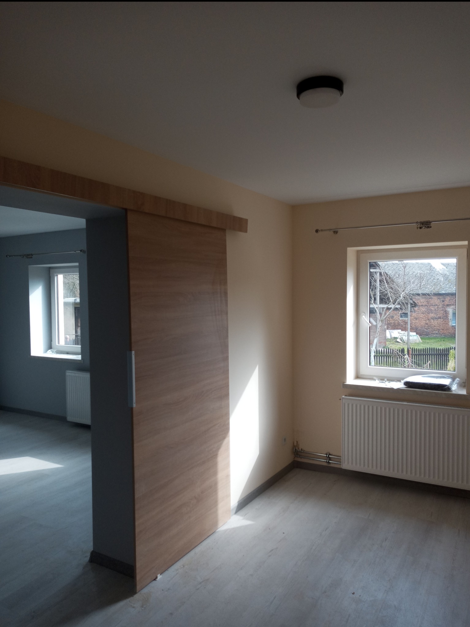 Wnętrze pokoju po remoncie z przesuwnymi drzwiami z okleiną drewnopodobną, grzejnikiem pod oknem i świeżo pomalowanymi ścianami w kolorze kremowym.