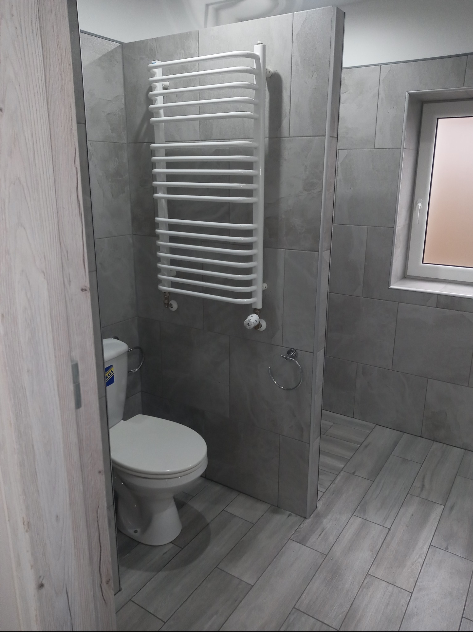 Wykończona łazienka w szarościach: płytki imitujące drewno na podłodze i ścianach, biała toaleta i grzejnik drabinkowy, widok zza uchylonych drzwi.