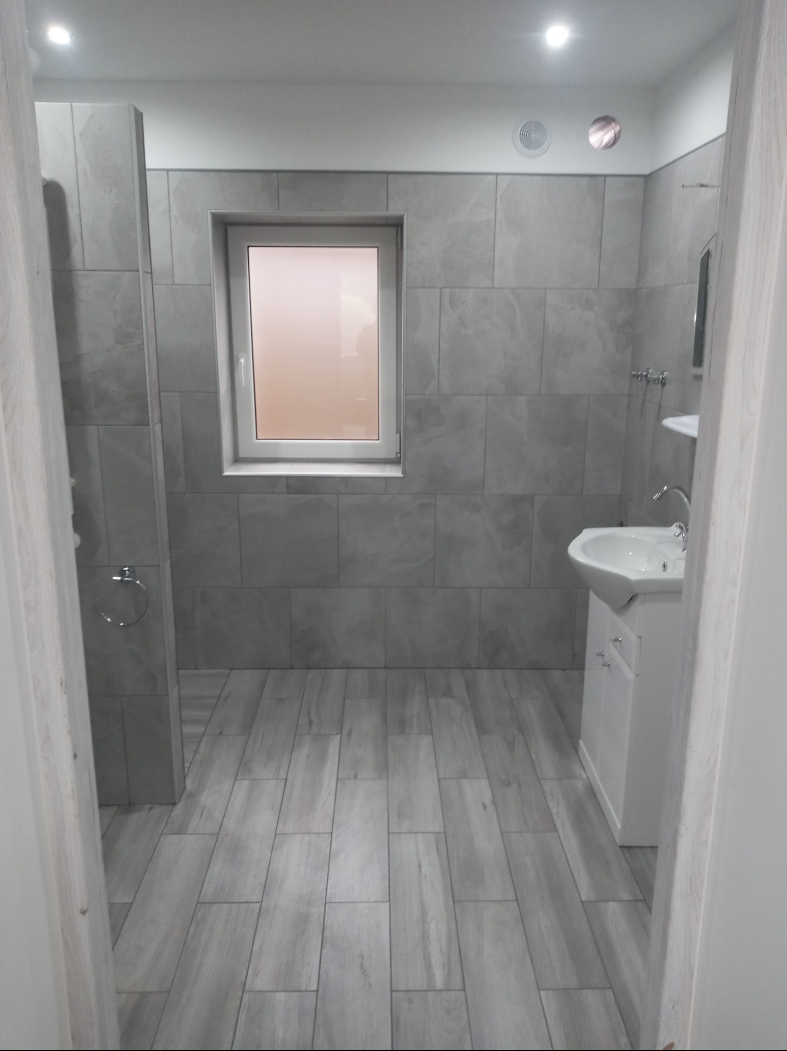 Wykończona łazienka w szarościach: płytki imitujące kamień na ścianach i podłodze, biała umywalka z szafką, okno z rozmytym widokiem, minimalistyczne oświetlenie sufitowe.