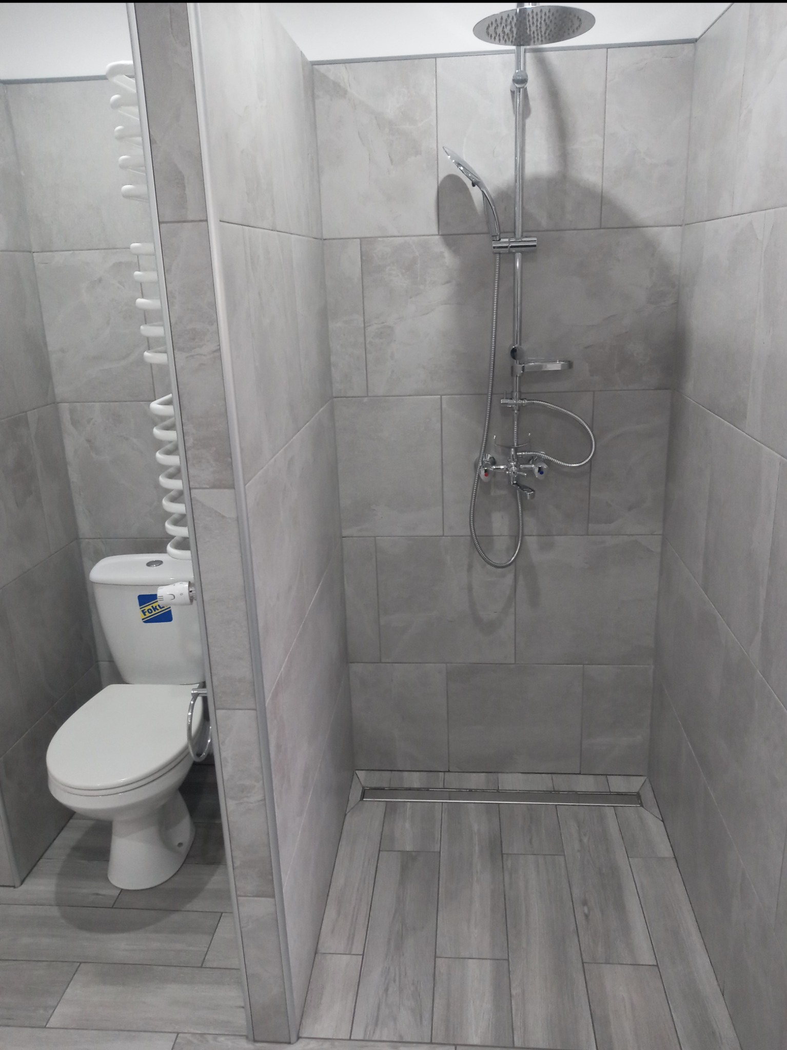 Wykończona łazienka w odcieniach szarości, z kabiną prysznicową typu walk-in, odpływem liniowym w podłodze wyłożonej płytkami imitującymi drewno oraz toaletą z grzejnikiem drabinkowym.