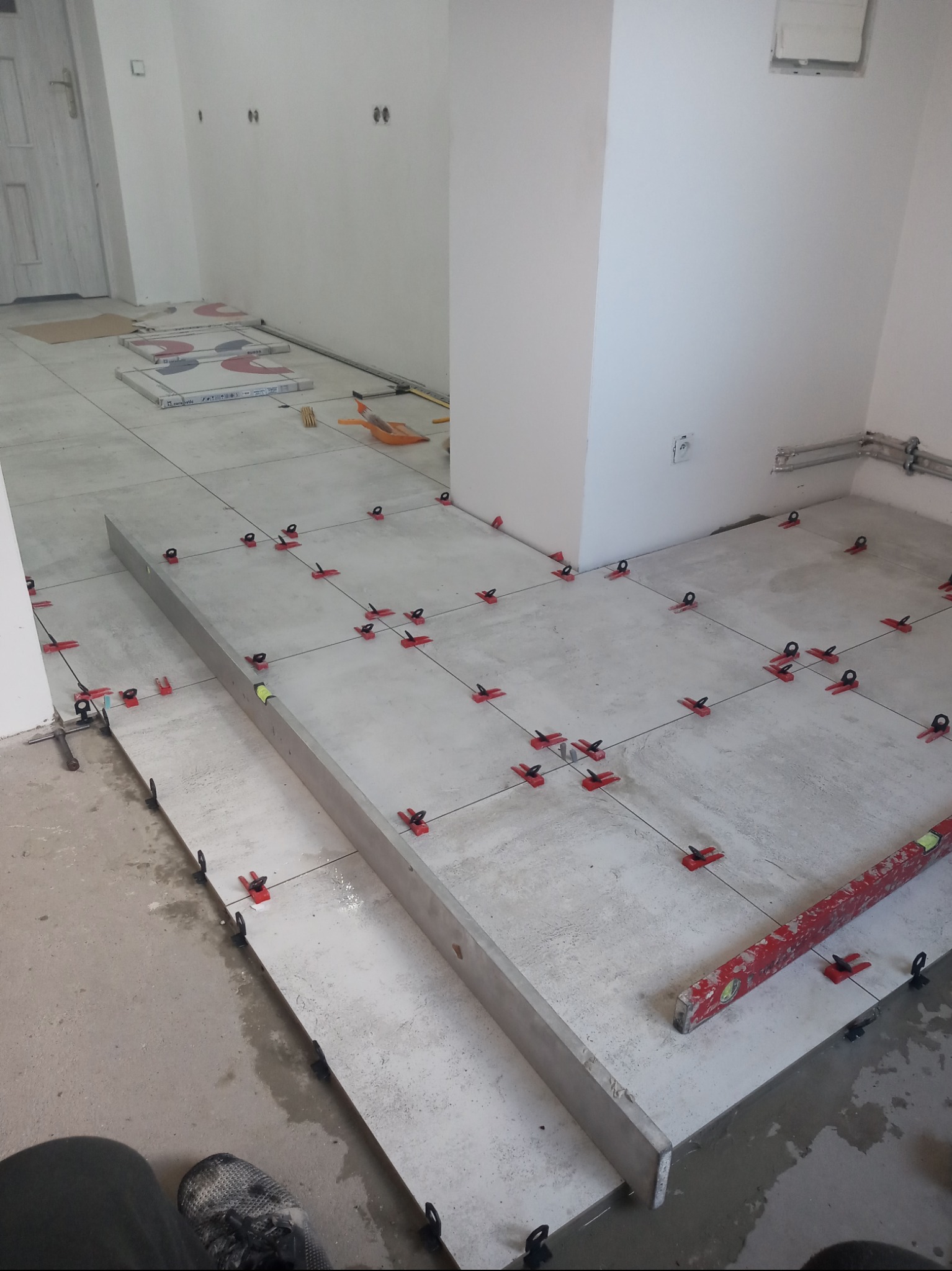 Układanie szarych płytek podłogowych z użyciem systemu poziomowania płytek, widoczna poziomica i puszki elektryczne w ścianie.