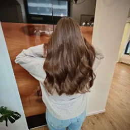 Długie, brązowe, falowane włosy kobiety, widok z tyłu, opierającej ręce na błyszczącej, drewnianej szafce w jasnym pomieszczeniu.