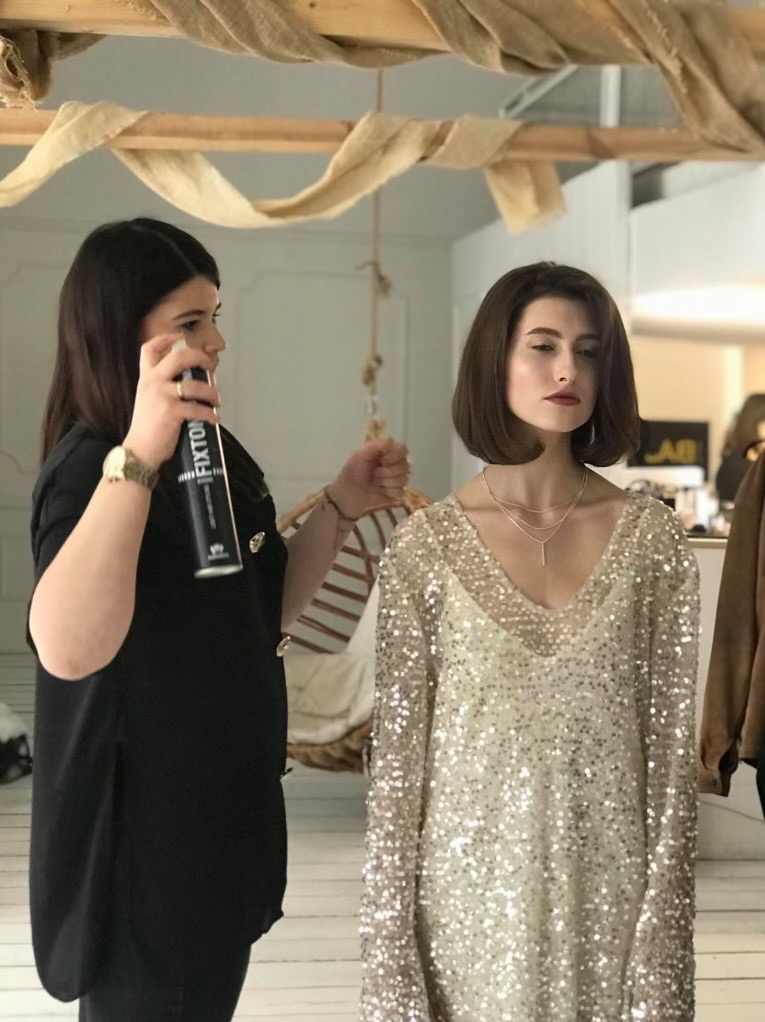 Stylistka fryzur utrwala lakierem krótkie, ciemne włosy modelki ubranej w cekinową sukienkę; widoczny fragment scenografii z drewnianymi belkami i beżową tkaniną.