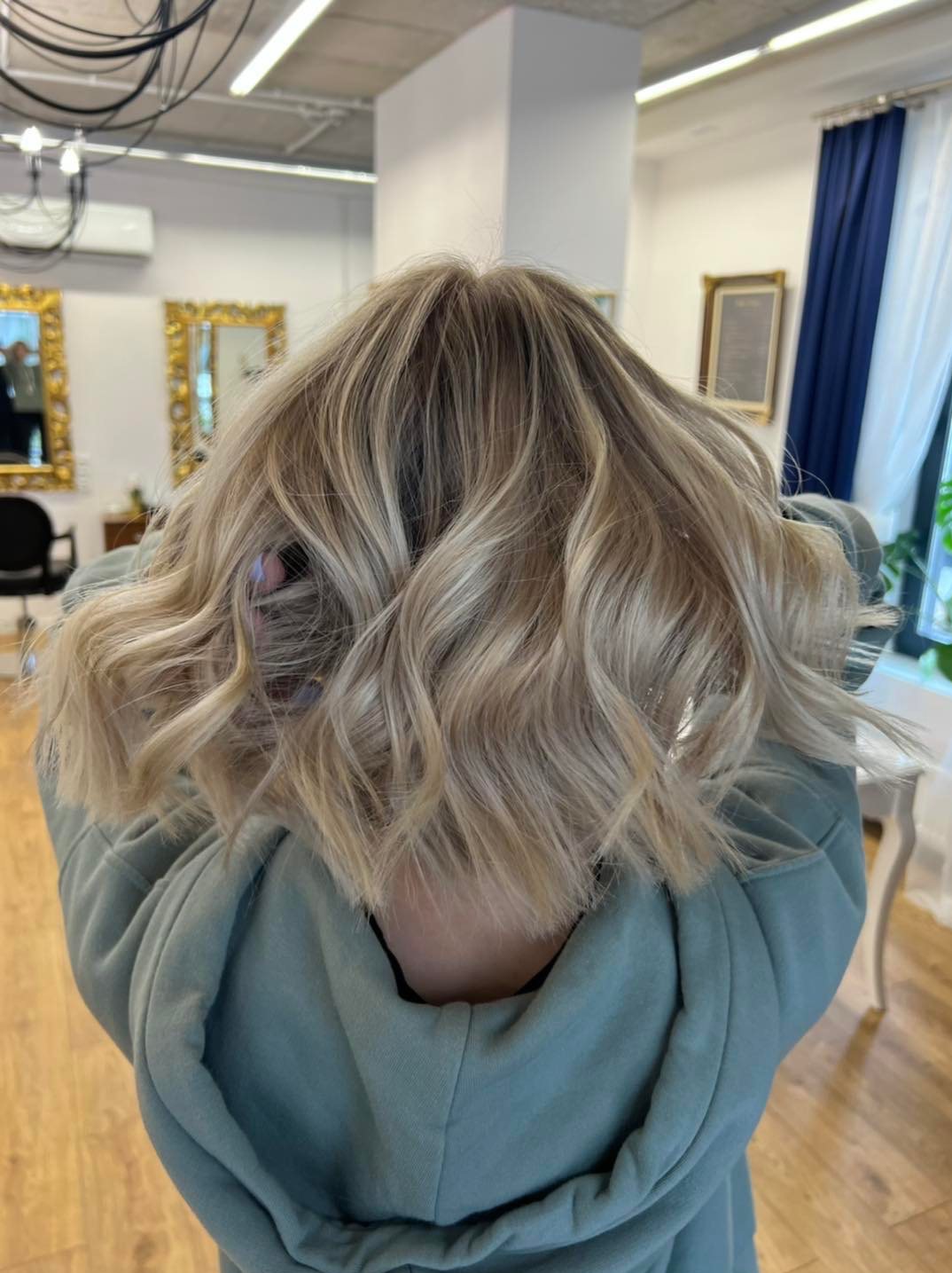 Krótkie, falowane włosy w odcieniach blondu z ciemniejszym odrostem, widok z tyłu, w tle wnętrze salonu z lustrami i zasłonami.