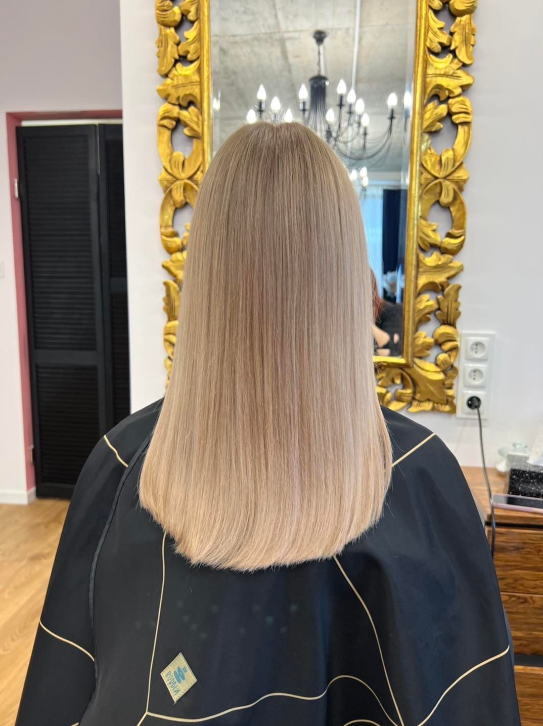 Proste, blond włosy po koloryzacji i strzyżeniu, widok z tyłu na tle lustra w złotej ramie i czarnej peleryny ochronnej.