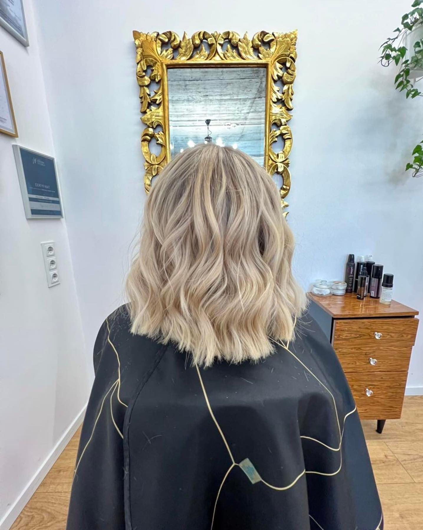 Blond włosy po koloryzacji i strzyżeniu, widoczne z tyłu, na tle lustra w złotej, zdobionej ramie i komody z kosmetykami w salonie.