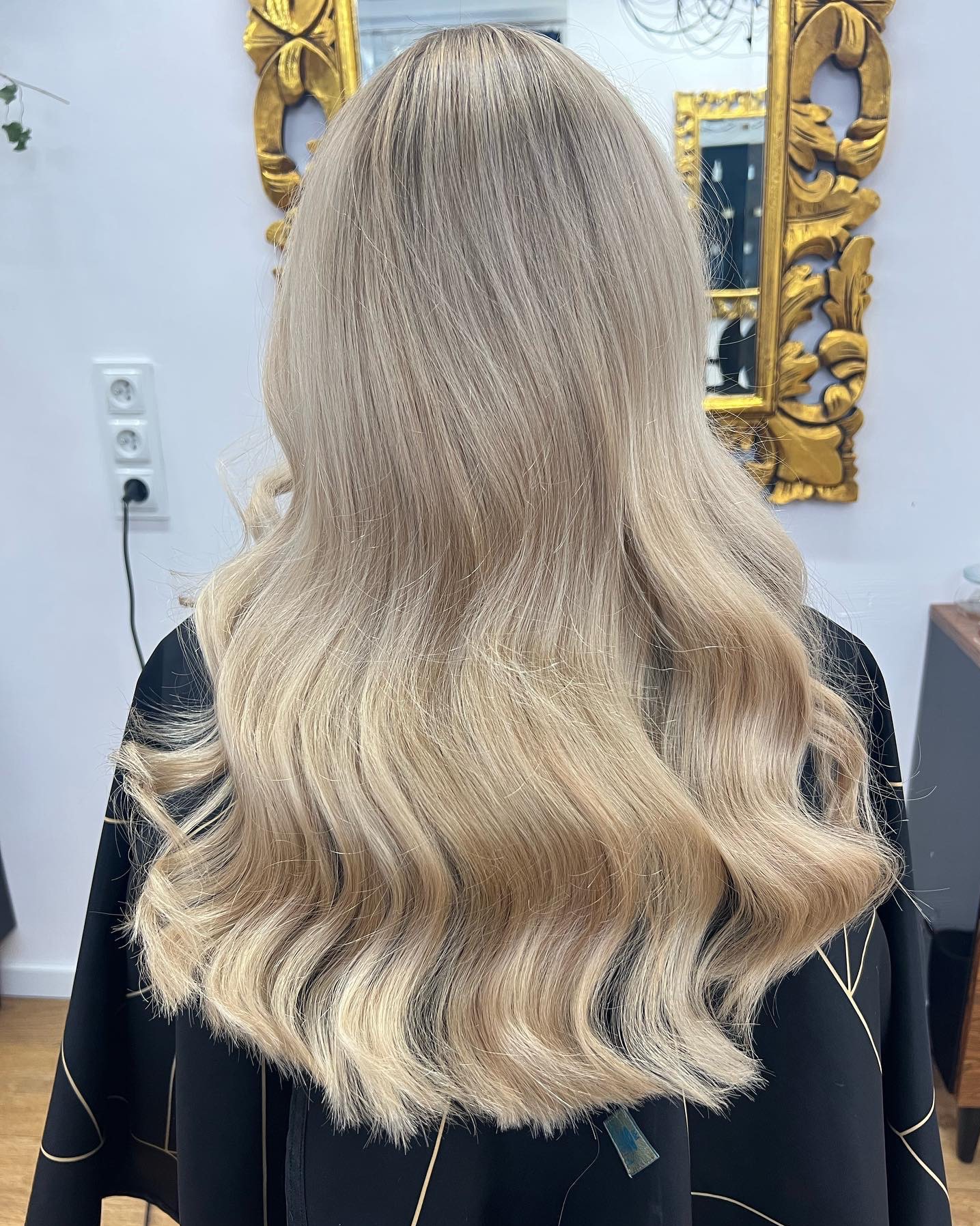 Długie, blond włosy z delikatnymi falami, widok z tyłu, czarna peleryna z geometrycznym wzorem, złote lustro w ozdobnej ramie w tle.