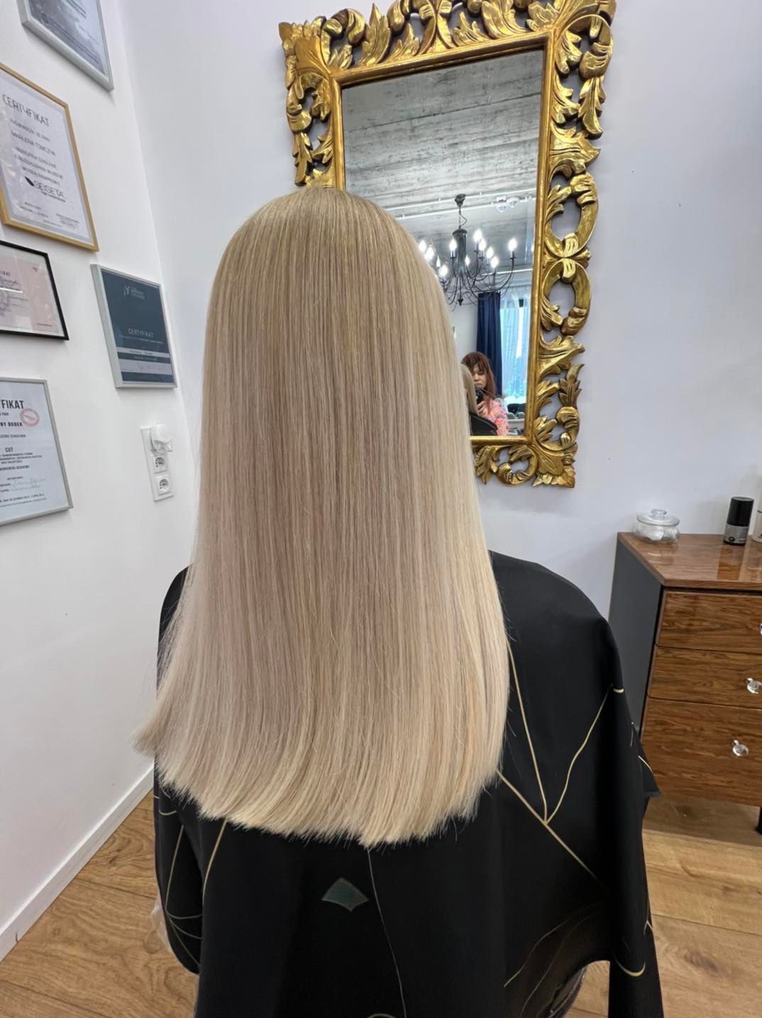 Tył głowy kobiety z długimi, prostymi, blond włosami po koloryzacji, widoczne certyfikaty na ścianie i odbicie fryzjerki w ozdobnym lustrze.