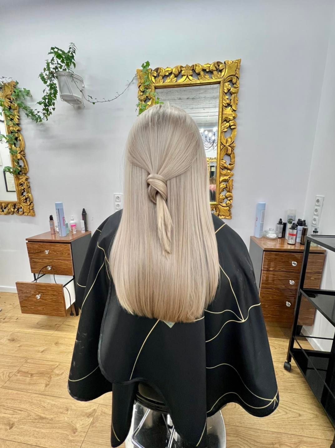 Jasne, proste włosy z delikatnym węzłem z tyłu głowy, widok z tyłu na tle salonu z lustrami w złotych ramach.